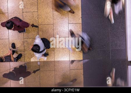 Vue aérienne des gens qui traversent le centre commercial. Le mouvement flou contraste avec un groupe stationnaire de personnages conversant. Scène représente un environnement urbain animé. Banque D'Images