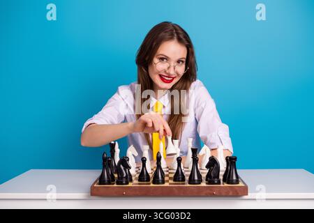 Jeune femme dans des lunettes jouant aux échecs sur une planche de bois tout en souriant avec confiance sur un fond bleu dégradé Banque D'Images