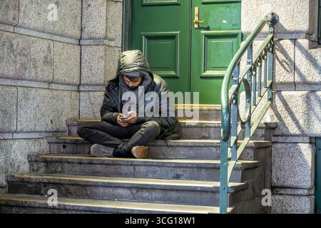 Un homme en sweat à capuche s'assoit sur des marches à l'aide d'un smartphone dans un bâtiment le long de Flinders Street à Melbourne, en Australie. Banque D'Images