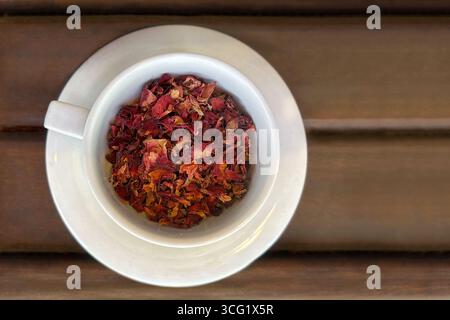 Une vue de haut en bas d'une tasse en céramique blanche remplie de pétales de rose séchés, placée sur une soucoupe assortie sur une surface en bois. Concept de beauté naturelle, herbe Banque D'Images