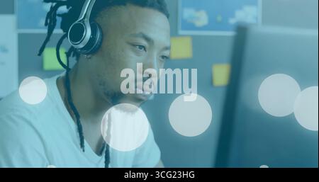 Homme regardant portant un casque supra-aural noir assis à un bureau, avec un tableau de notes autocollantes et un moniteur Banque D'Images