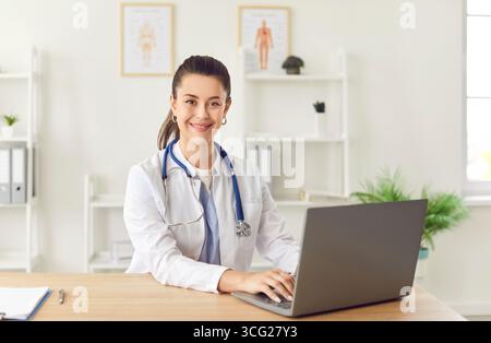 Portrait de jeune médecin féminin souriant confiant en uniforme médical blanc assis au bureau de travail Banque D'Images