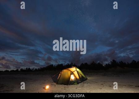 Une tente de camping confortable et éclairée brille chaleureusement sur une plage de sable éloignée sous un ciel nocturne dramatique avec des étoiles et des nuages. Banque D'Images