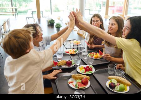 Un groupe animé d'étudiants dégustant des fruits frais et des collations ensemble pendant le déjeuner dans une cafétéria de l'école propre et lumineuse. La scène met en vedette Friendshi Banque D'Images