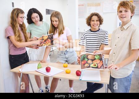 Un groupe d'adolescents s'est réuni autour d'une table dans une salle de classe d'école présentant des options alimentaires saines, tenant des photos de repas et s'engageant dans une discussion Banque D'Images