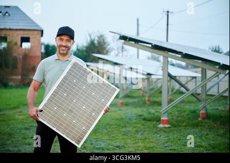 L'homme tient le panneau solaire tout en se tenant debout dans la ferme solaire. Joyeux gars portant une casquette noire et un polo vert clair. Arrière-plan avec des rangées de panneaux solaires et un vieux bâtiment en briques, placé contre un ciel clair et nocturne. Banque D'Images