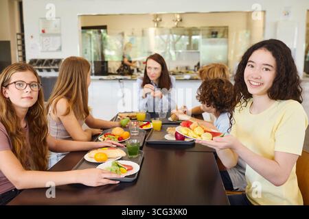 Un groupe diversifié d'étudiants se réunissent dans une cafétéria de l'école lumineuse, appréciant une variété de repas sains avec des produits frais, du jus et de la conversation, refl Banque D'Images
