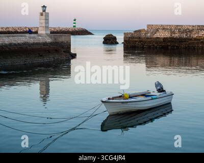 Bateau de pêche blanc amarré dans l'eau calme près d'une jetée de pierre avec phare au coucher du soleil. Lagos, Algarve, Portugal, 13 mai 2017. Banque D'Images