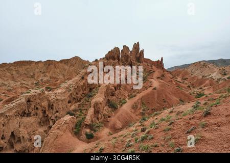 Formations rocheuses de grès rouge à Skazka, Fairytale Canyon, Kirghizistan, avec des falaises escarpées, des crêtes érodées, et végétation désertique clairsemée sous un nuageux Banque D'Images