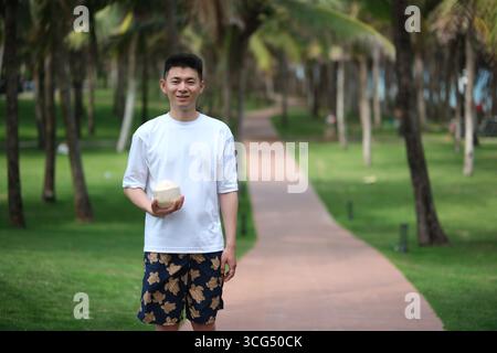 Jeune homme asiatique souriant avec de la noix de coco sur le chemin bordé de palmiers à Sanya, Hainan, Chine Banque D'Images