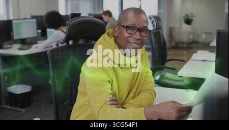 Homme portant un sweat à capuche jaune et des lunettes tapant à l'ordinateur dans le bureau ouvert, superposition de grille numérique Banque D'Images