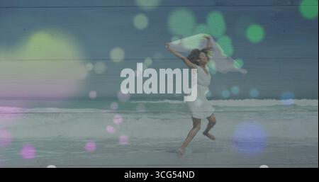 Femme en robe blanche courant le long de la plage tenant une écharpe blanche, avec des vagues de l'océan et des superpositions bokeh Banque D'Images