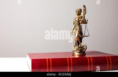 Statue dorée de la déesse de la justice tenant des écailles sur un livre marron qui est sur une table blanche sur un fond gris Banque D'Images