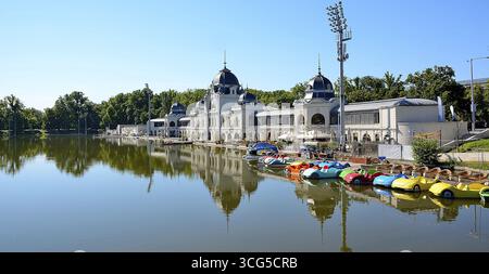 Les bateaux à aubes colorés sont alignés sur la rive d'un lac, avec un grand bâtiment et des arbres luxuriants en arrière-plan, reflétant dans l'eau calme de cit Banque D'Images