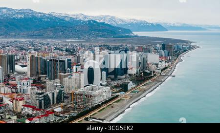 Panorama aérien révélant le paysage urbain de batoumi, avec gratte-ciel contemporains, hôtels de luxe, complexes résidentiels bordant la côte de la mer noire, sommets montagneux enneigés encadrant le paysage urbain Banque D'Images