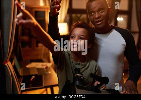 Père noir moderne et son fils passant la soirée ensemble à la maison s'amusant à regarder les étoiles à l'aide d'un télescope Banque D'Images