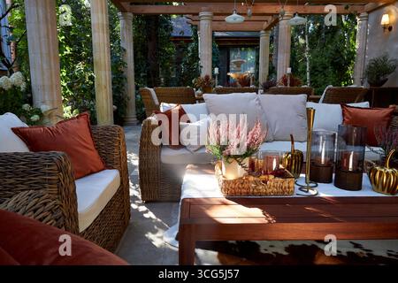 Coin salon extérieur élégant avec fauteuils en osier, coussins blancs et oreillers orange. Table en bois décorée de bougies, de plantes et de détails d'automne Banque D'Images