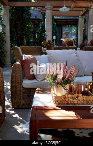 Coin salon extérieur élégant avec fauteuils en osier, coussins blancs et oreillers orange. Table en bois décorée de bougies, de plantes et de détails d'automne Banque D'Images