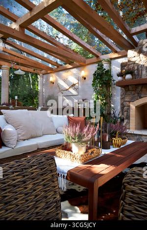 Coin salon extérieur élégant avec fauteuils en osier, coussins blancs et oreillers orange. Table en bois décorée de bougies, de plantes et de détails d'automne Banque D'Images