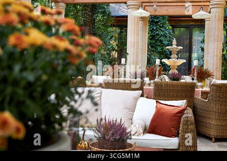 Coin salon extérieur élégant avec fauteuils en osier, coussins blancs et oreillers orange. Table en bois décorée de bougies, de plantes et de détails d'automne Banque D'Images