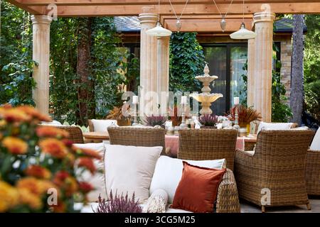 Coin salon extérieur élégant avec fauteuils en osier, coussins blancs et oreillers orange. Table en bois décorée de bougies, de plantes et de détails d'automne Banque D'Images