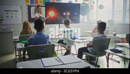 Élèves de l'école primaire étudiant l'information de base sur le système solaire et les planètes dans la salle de classe moderne. Enseignante éduquant Smart diverse Kids pendant la leçon d'astronomie à l'aide de l'écran numérique. Banque D'Images
