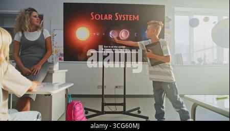 Garçon d'école élémentaire avec ordinateur tablette donnant présentation sur le système solaire et les planètes devant les camarades de classe et l'enseignant à l'aide de l'écran numérique. Smart Kids étudier l'astronomie spatiale en classe. Banque D'Images
