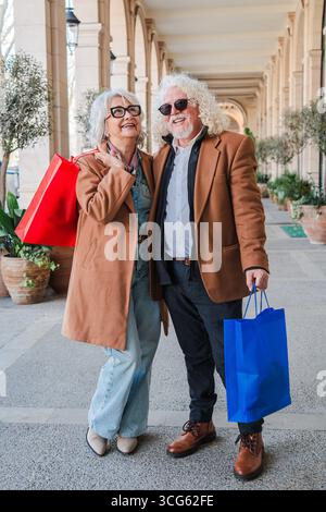 Un couple de personnes âgées joyeux s'engageant joyeusement dans une aventure de shopping délicieuse tout en se promenant et tenant des sacs vibrants remplis de trouvailles passionnantes Banque D'Images
