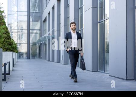 Un homme professionnel en costume sombre marche en toute confiance à l'extérieur à côté d'une façade en verre contemporaine. Banque D'Images