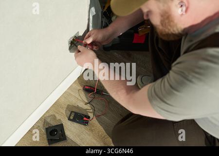 Homme d'âge moyen caucasien agenouillé sur un plancher en bois testant la prise électrique avec un multimètre numérique, tenant les sondes près de la prise, effectuant des travaux de maintenance ou de réparation Banque D'Images
