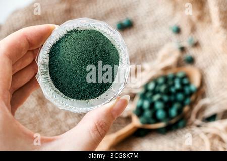Main femelle tient la poudre de spiruline sur un fond de toile de jute et une cuillère avec des comprimés d'algues vertes Banque D'Images