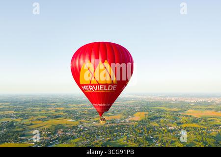 Sint-Martens-Latem, Belgique - 25 août 2025 : vue aérienne d'une montgolfière rouge survolant le paysage verdoyant et luxuriant, un phare contre le ciel bleu et doux. Banque D'Images