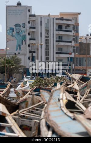 Dakar, Sénégal - 12 mai 2025 : vue sur les bateaux de pêche dynamiques de Soumbédioune reposant à terre sur un fond de bâtiments modernes, créant un contraste captivant. Banque D'Images