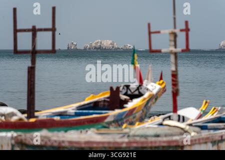 Dakar, Sénégal - 12 mai 2025 : vue de bateaux de pêche dynamiques reposant sur la côte sablonneuse, leurs coques colorées contrastant avec les îles lointaines et brumeuses et la mer calme et grise. Banque D'Images