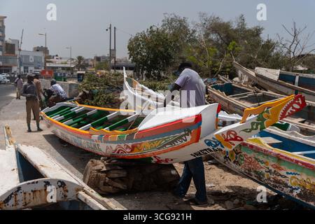 Dakar, Sénégal - 12 mai 2025 : vue de bateaux aux couleurs vives qui reposent près de la plage de Soumbédioune, où les artisans travaillent méticuleusement, mêlant tradition et pulsation vibrante de la vie sénégalaise. Banque D'Images