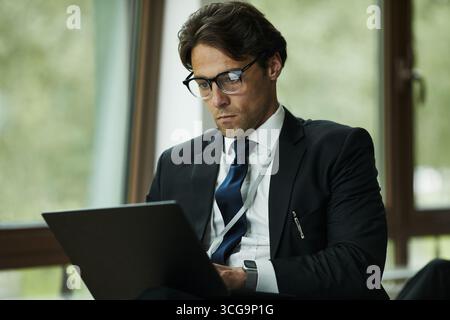 Jeune homme caucasien portant un costume assis à l'intérieur travaillant sur un ordinateur portable avec une expression ciblée, cheveux bruns courts et lunettes visibles, grande fenêtre en arrière-plan Banque D'Images