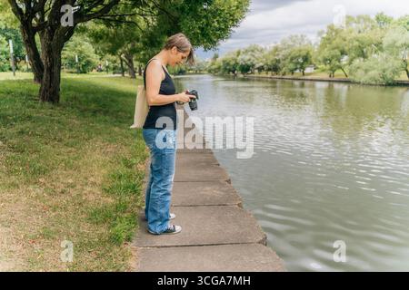 Femme des années 40 examinant des photos sur la caméra par la rive urbaine de la rivière avec sac fourre-tout Banque D'Images