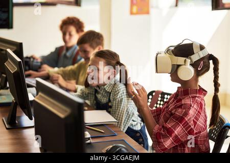 Groupe d'enfants caucasiens et noirs assis à des bureaux à l'aide d'ordinateurs en classe, fille portant un casque de réalité virtuelle interagissant avec le contenu numérique, camarades de classe regardant et souriant Banque D'Images
