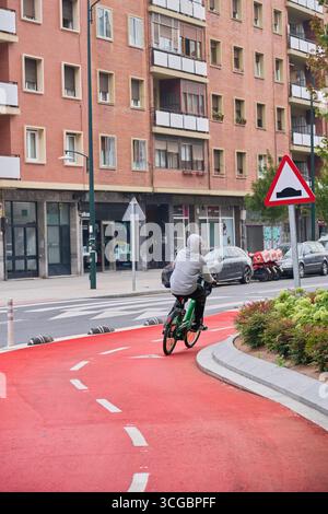 Adolescent conduisant un vélo vert sur une piste cyclable rouge vibrante dans un cadre urbain, promouvant le transport durable Banque D'Images