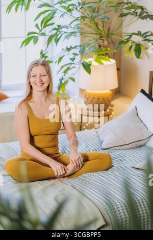 Jeune femme vêtue de vêtements de sport jaune vif pratiquant le yoga et la méditation sur un lit confortable dans une chambre lumineuse et accueillante ornée de plantes vertes luxuriantes et d'une lampe élégante Banque D'Images