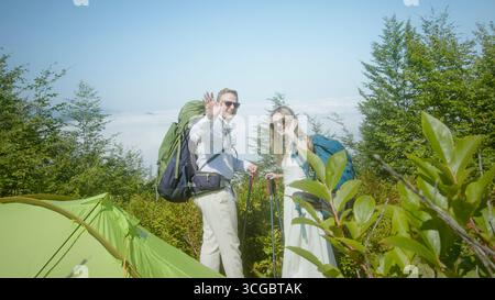 Couple heureux avec des sacs à dos ondulant près d'une tente verte dans une forêt de montagne luxuriante, profitant du voyage d'aventure et de la randonnée estivale. Banque D'Images