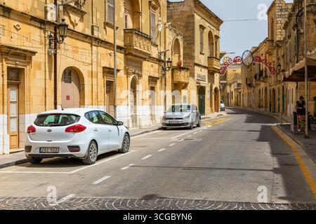 Gozo, Malte – 23 juin 2021 : vue sur la rue avec des bâtiments traditionnels en calcaire, des décorations festives et des voitures garées. Banque D'Images