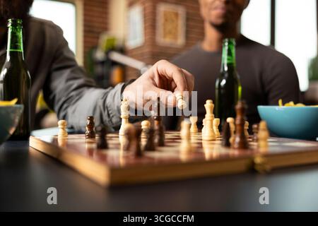 Gros plan d'un match d'échecs occasionnel en cours alors que le joueur masculin caucasien déplace une pièce. Échiquier sur la table, entouré de boissons et d'en-cas tandis que les amis profitent d'une séance de jeu de société intérieure détendue. Banque D'Images