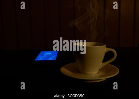 Une tasse de café chaude avec de la vapeur qui monte, éclairée par une lumière chaude, à côté d'un téléphone qui vérifie la météo. Un concept pour un matin confortable, slow living, rel Banque D'Images