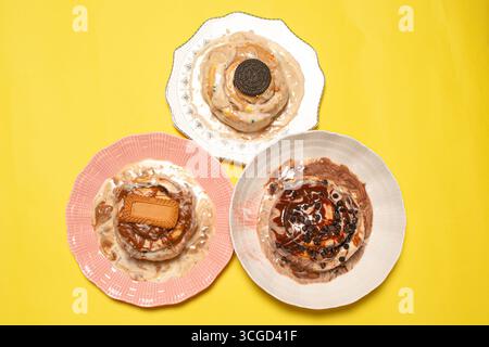Vue de haut en bas d'un assortiment de trois petits pains gourmands à la cannelle sur des assiettes colorées avec différentes garnitures sucrées sur un fond jaune vif solide Banque D'Images