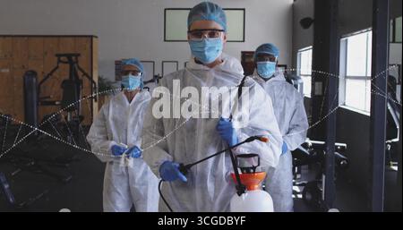 Désinfectant travailleur portant des combinaisons blanches, des filets à cheveux, des lunettes et des gants dans le gymnase avec une baguette de pulvérisateur Banque D'Images