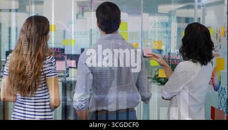 Collègues collaborateurs esquissant des diagrammes sur un mur de verre dans le bureau, avec des notes adhésives colorées Banque D'Images