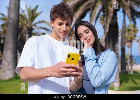 Couple sur la pelouse en bord de mer partageant un smartphone jaune, petit ami pointant vers l'écran Banque D'Images