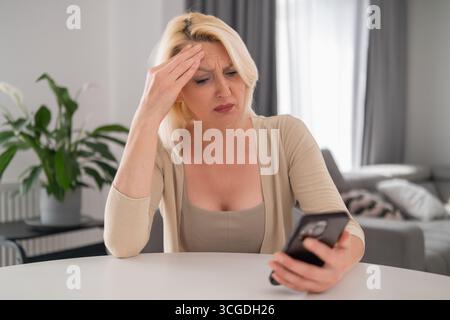 Femme blonde d'âge moyen assise seule dans un salon confortable, tenant un smartphone et regardant l'écran avec un regard pensif et inquiet, reflétant le stress et l'incertitude de la vie moderne Banque D'Images