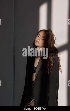 Femme aux cheveux longs porte un blazer noir et un haut, debout contre un mur baigné de lumière douce du soleil, exsudant le calme et l'élégance dans un sp minimaliste Banque D'Images